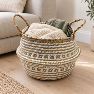 Könnte beinhalten: Ein runder geflochtener Korb mit einem geometrischen Muster in Weiß und Naturbeige sowie zwei geflochtenen Griffen. Der Korb ist mit einer beigen, strukturierten Decke und einem gefalteten olivgrünen Stoff gefüllt und hat eine breite Basis.