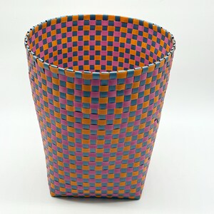 May include: A cylindrical wastebasket constructed from woven plastic strips in a repeating checkerboard pattern. The basket features alternating colors of bright orange, vibrant pink, and slate blue, creating a uniform grid design throughout.