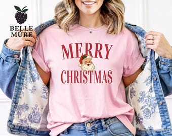 May include: A light pink t-shirt with the words "MERRY CHRISTMAS" in red, along with a Santa Claus graphic. The shirt is part of a Christmas-themed collection. The model is wearing a denim jacket.
