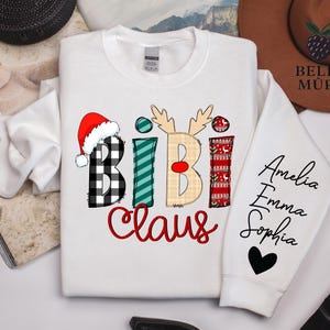 May include: White sweatshirt with the text "Bibi Claus" in a festive design. The letter "B" features a black and white buffalo plaid pattern, and the letter "I" is decorated with green stripes. The sleeve displays the names Amelia, Emma, and Sophia.