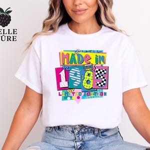 May include: White t-shirt with a colorful graphic that reads "MADE IN 1985 LIMITED EDITION." The design features the number 1985 in a retro style with bright colors and a checkered pattern. The shirt has a classic crew neck.