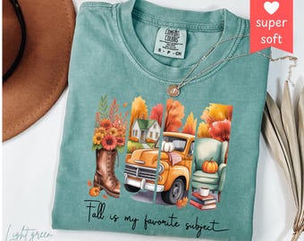 Puede incluir: Una camiseta verde claro con un estampado gráfico de una bota marrón llena de flores, un camión naranja, una casa y un sillón con calabazas. El texto "Fall is my favorite subject" está impreso debajo del gráfico. Una etiqueta dice "Comfort Colors".