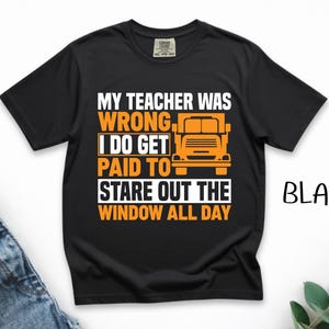 Op de afbeelding: Een zwart t-shirt met korte mouwen en een grafische print in oranje en wit. De tekst luidt: MY TEACHER WAS WRONG I DO GET PAID TO STARE OUT THE WINDOW ALL DAY. Een oranje icoon van een vrachtwagen staat aan de rechterkant van de tekst.