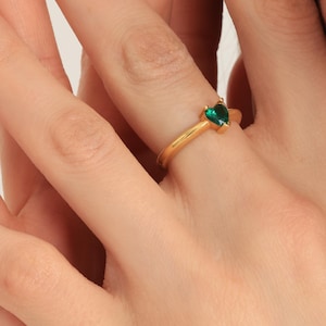 May include: A gold ring featuring a heart-shaped, emerald-green gemstone. The ring has a delicate band with a textured design. The ring is shown on a person's finger.
