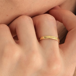 May include: A gold-colored ring with the word "Lucky" engraved on the band. The ring is worn on a finger. The image is a close-up of the hand and face.