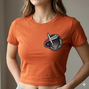 May include: Orange crop top t-shirt with a circular patch featuring a rocket design and the text "PROJECT HAIL MARY". The patch has a black border with white and red accents. The shirt has short sleeves and a crew neck.