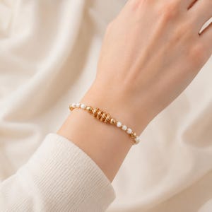 May include: Gold beaded bracelet with white pearl accents. The bracelet features a central design of gold rings and beads. The bracelet is worn on a wrist against a soft, off-white background.