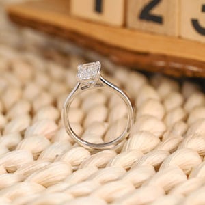 Peut inclure: Une bague de fiançailles en argent avec un diamant de taille carrée. La bague est posée sur une surface beige tressée. Le corps de la bague est fin et poli, et le diamant est clair et scintillant.