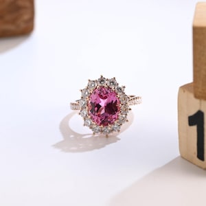 May include: A rose gold ring featuring a large, oval-cut pink gemstone surrounded by smaller, clear gemstones. The ring's band is also adorned with small, clear gemstones, creating a halo effect. The ring is set against a white background.