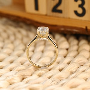 Puede incluir: Un anillo de compromiso dorado con una gran piedra central transparente. El anillo tiene una banda delicada y piedras de acento más pequeñas engastadas en el puente. Se muestra sobre una superficie beige tejida.