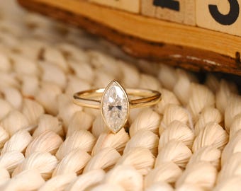 Könnte beinhalten: Ein goldener Ring mit einem Diamanten im Marquiseschliff. Der Diamant ist in einer Zargenfassung gefasst und das Band ist dünn. Der Ring wird auf einer gewebten Oberfläche präsentiert, mit einem Holzblockkalender im Hintergrund.