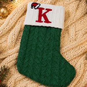 May include: A green cable-knit Christmas stocking featuring a white ribbed cuff with a red embroidered letter K and a small Santa hat icon. The stocking is displayed on a textured cream-colored fabric surface surrounded by pine branches and ornaments.