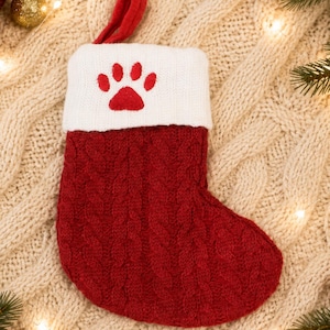 May include: A red cable-knit Christmas stocking featuring a white cuff with a red embroidered paw print design. The stocking includes a red fabric loop at the top for hanging and is constructed with a textured knit pattern throughout the body.