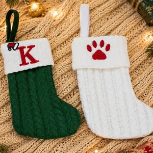 May include: Two cable-knit Christmas stockings. One is dark green with a white cuff featuring a red letter K and a small Santa hat. The other is white with a white cuff featuring a red embroidered paw print. Both have matching hanging loops.