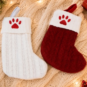 May include: Two knit Christmas stockings, one white and one deep red, each featuring a white cuff embroidered with a red paw print. The stockings have a cable knit texture and a hanging loop at the top, designed for holiday decoration.
