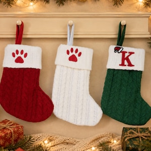 May include: Three cable-knit Christmas stockings hanging from a wooden rack. The left is red with a white cuff and red paw print. The center is white with a white cuff and red paw print. The right is green with a white cuff and red letter K.