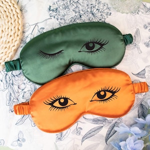 May include: Two satin eye masks, one green and one orange, each featuring a black eye design with long eyelashes. The masks have elastic bands for a comfortable fit. The background is a floral patterned fabric.
