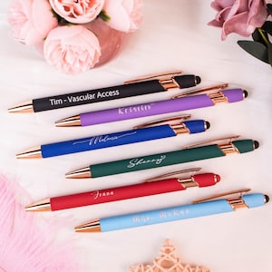 May include: A selection of personalised pens in colours such as black, purple, blue, green, red, and light blue. Each pen has gold-coloured tips and accents, with names or text printed on them. The pens are arranged on a white surface.