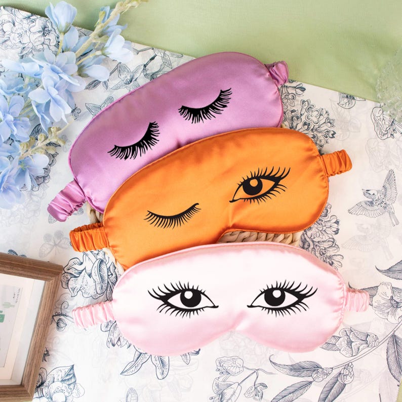 May include: Three satin sleep masks in shades of pink, orange, and purple, each adorned with a black eye and lash design. The masks have elastic bands for a comfortable fit. The masks are arranged on a floral patterned surface.