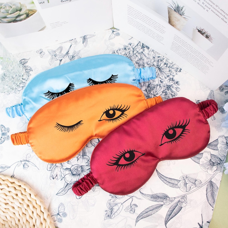 May include: Three satin sleep masks in blue, orange, and burgundy, each featuring a black eyelash design. The masks have elastic bands for a comfortable fit and are displayed on a floral patterned surface.
