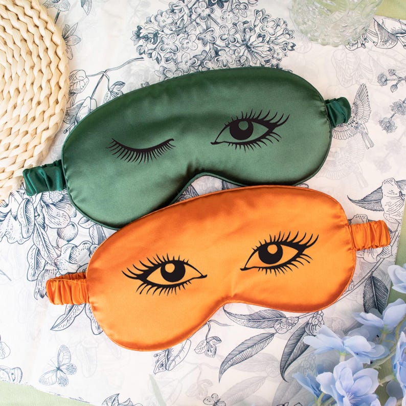 May include: Two satin eye masks, one green and one orange, each featuring a black eye design with long eyelashes. The masks have elastic bands for a comfortable fit. The background is a floral patterned fabric.