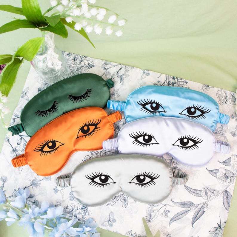 May include: A collection of satin sleep masks in various colors, including green, orange, blue, white, and silver. Each mask features a stylized eye design with long eyelashes. The masks are arranged on a floral patterned fabric.
