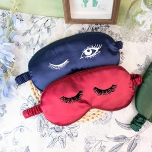 May include: Two satin sleep masks. One is navy blue with a white eye design, and the other is red with black eyelashes. The masks are on a woven mat, with a floral patterned background.