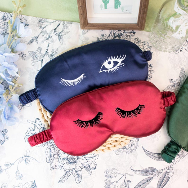 May include: Two satin sleep masks. One is navy blue with a white eye design, and the other is red with black eyelashes. The masks are on a woven mat, with a floral patterned background.