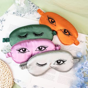 May include: Four satin sleep masks in shades of green, orange, pink, and silver. Each mask features a black eye design with long eyelashes. The masks are laying on a white surface with floral accents.
