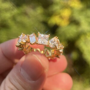 Puede incluir: Un anillo dorado con una serie de piedras preciosas claras y brillantes en varias formas, incluyendo cortes cuadrados, esmeralda y pera. El anillo se sostiene contra un fondo verde borroso, resaltando el diseño intrincado y el brillo de las piedras.
