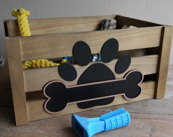 May include: A wooden pet toy storage crate with a paw print and bone design. The crate is filled with various dog toys, including a yellow rope toy and a blue chew toy. The crate is made of light brown wood.