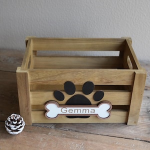 Peut inclure: Caisse en bois avec une empreinte de patte et un motif d'os. L'os porte le nom "Gemma". La caisse est en bois brun clair et de forme rectangulaire. Une pomme de pin est dans le coin inférieur gauche.