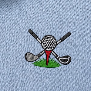 May include: Light blue polo shirt with a golf-themed embroidered design. The design features a golf ball on a red tee, crossed golf clubs, and green grass. The shirt has a button-down collar.