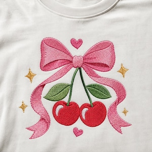 Puede incluir: Una camiseta blanca con un diseño bordado de dos cerezas rojas con hojas verdes, unidas por un gran lazo rosa. El diseño incluye pequeños corazones rosas encima y debajo de las cerezas, junto con cuatro estrellas doradas bordadas.