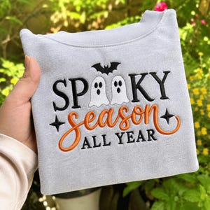 Puede incluir: Una sudadera gris claro de cuello redondo con bordados en negro y naranja. El diseño incluye dos fantasmas blancos, un murciélago negro y el texto "SPOOKY season ALL YEAR" en una mezcla de letras negras y cursiva naranja con detalles de estrellas.