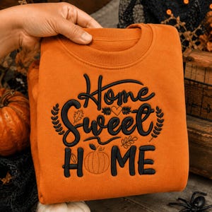 May include: An orange crewneck sweatshirt featuring black embroidered text that reads Home Sweet HOME. The design includes a small embroidered heart, leaf motifs, and a pumpkin outline integrated into the lettering on the front of the garment.