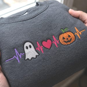 May include: A dark gray crewneck sweatshirt featuring an embroidered heartbeat line design. The pattern includes a white ghost, a pink heart, and an orange jack-o'-lantern pumpkin, with the heartbeat line transitioning through purple, pink, and orange.