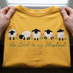 May include: A mustard yellow t-shirt featuring an embroidered row of five white sheep with black faces and legs, interspersed with small pink flowers and green grass. Below the sheep, the text "the Lord is my Shepherd" is embroidered in green.