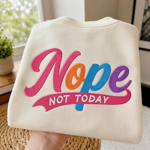 Könnte beinhalten: Ein cremefarbenes Sweatshirt mit Rundhalsausschnitt und großen, bunten Stickereien auf der Vorderseite. Das Wort Nope ist in Pink, Orange, Blau und Lila gestickt, mit dem in Weiß auf einem pinken Schweif gestickten Text NOT TODAY.