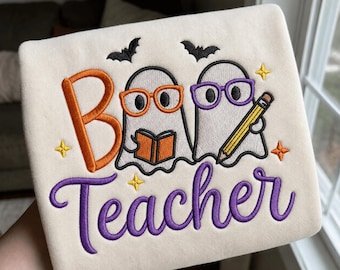Puede incluir: Una sudadera color crema con un diseño bordado de dos fantasmas. Un fantasma lleva gafas naranjas y sostiene un libro naranja, mientras que el otro lleva gafas moradas y sostiene un lápiz amarillo. El texto dice "Boo Teacher" en naranja y morado.