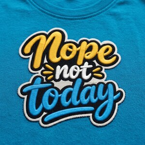 May include: A bright blue t-shirt featuring a large, multi-colored embroidered patch on the chest. The patch displays the text "Nope not today" in a stylized, layered font with yellow, white, and light blue lettering outlined in black.