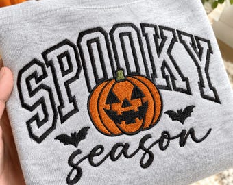 Puede incluir: Una sudadera gris claro con texto bordado que dice SPOOKY en una fuente universitaria negra y season en letra cursiva negra. Una calabaza naranja con rasgos faciales negros y dos pequeños murciélagos negros están centrados en el diseño.
