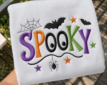 May include: A white sweatshirt featuring embroidered Halloween-themed designs. The word SPOOKY is stitched in purple, orange, black, and green. The design includes a spider web, two black bats, a hanging spider, and several small stars.