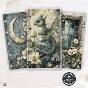 Peut inclure: Un ensemble de trois marque-pages rectangulaires pour agenda illustrés d'un dragon vert, d'un croissant de lune et d'une fenêtre avec des fleurs blanches. Les motifs incluent du texte manuscrit, de la dentelle et des textures de bois bleu.