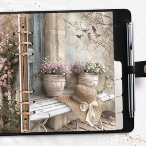 Könnte beinhalten: Ein schwarzer Planer mit einem floralen Einsatz, der einen Strohhut, Blumentöpfe und eine Bank zeigt. Ein silberner Stift ist angebracht. Der Planer hat goldfarbene Ringe.