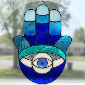 May include: A stained glass Hamsa hand with an evil eye design. The hand features shades of blue, teal, and white. The eye is in the center of the palm, with a blue pupil. The image has the text "IMAGI VERO" in the upper left corner.