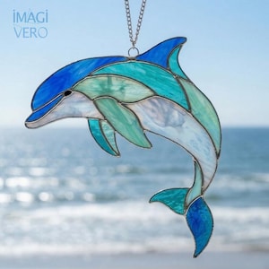 Könnte beinhalten: Glasfenster-Suncatcher in Form eines Delfins in Blau- und Türkistönen, mit einer silbernen Kette zum Aufhängen. Das Delfin-Design zeigt eine verspielte Pose, mit dem IMAGI VERO-Logo oben links. Der Hintergrund zeigt das Meer.
