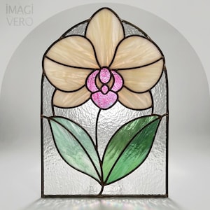 Pode incluir: Painel de vitral com um design de flor de orquídea. A flor tem pétalas creme com um centro rosa, sobre um fundo de vidro texturizado. Folhas verdes e uma moldura escura completam a obra de arte. A marca "IMAGI VERO" é visível.