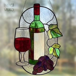 Op de afbeelding: Een ovaal glas-in-lood zonnevanger met een groene wijnfles, een glas gevuld met dieprode vloeistof, een tros paarse druiven en groene bladeren. De tekst IMAGI VERO is zichtbaar in de linkerbovenhoek op het heldere, getextureerde glas.