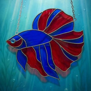 May include: Stained glass betta fish in shades of blue and red, with a small blue gem eye. The fish is suspended by a silver chain against a blue background with bubbles.